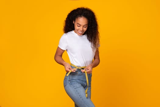 African American woman measures waist with tape smiling, yellow background
