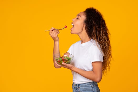 Side view of slender black lady with bowl of salad, eating healthy vegetable lunch, showcasing slimming meal, standing near copy space on vibrant yellow studio background. Nutrition concept
