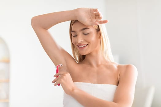 Attractive blonde lady using razor while shaving armpits in bathroom