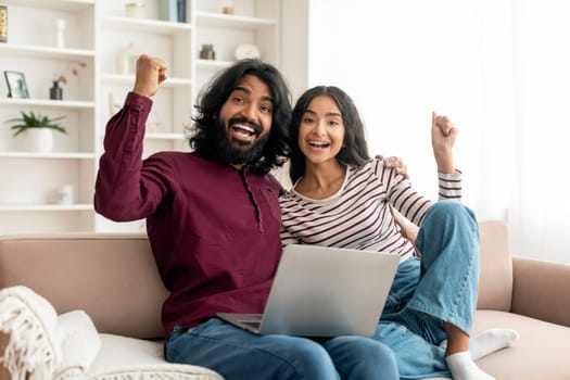 Emotional indian couple sitting on couch at home, using laptop