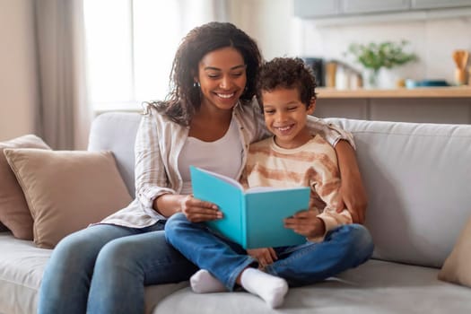 Cheerful black mother and her preteen son reading book together at home