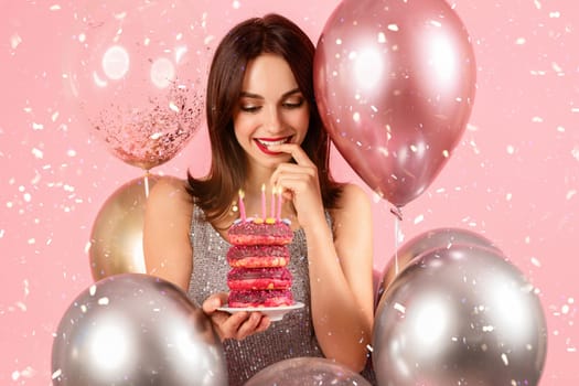 Charming woman with a secretive smile, playfully contemplating a bite of a stacked pink donut cake