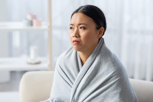 Depressed asian woman wrapped in blanket sitting on couch