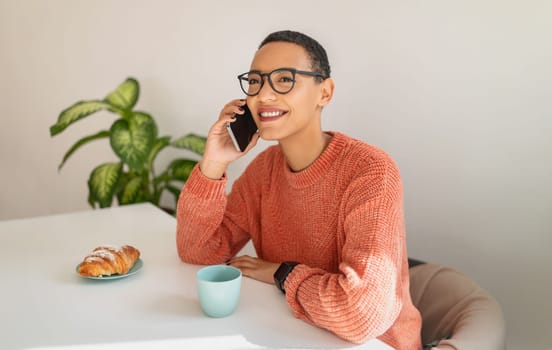 Happy millennial latin lady call by smartphone, drink cup of coffee, eat croissant