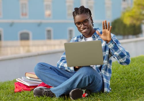 African student guy with laptop video calls waving hand outdoors