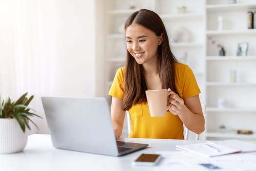 Young Smiling Asian Freelancer Woman Using Laptop And Drinking Coffee At Home