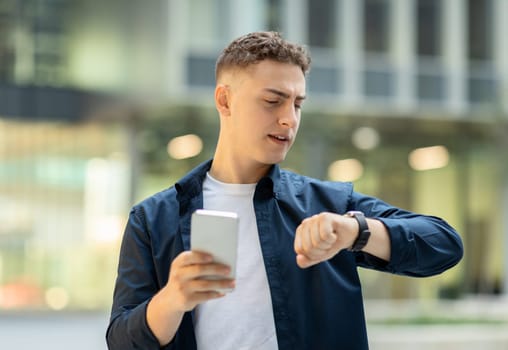 Busy young man with curly hair checking time on his smartwatch while holding a smartphone