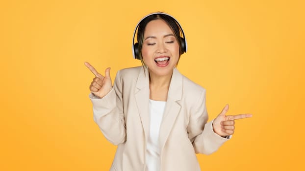 Happy millennial japanese lady in suit and wireless headphones has fun, singing song