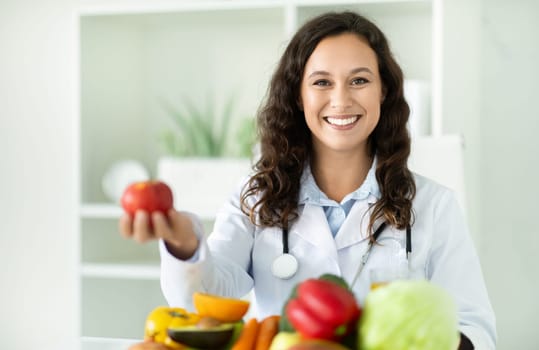 Young european woman dietitian holding apple, sitting at desk at clinic