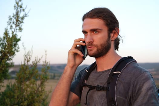 Male hiker with a backpack is talking on the phone in the countryside close up