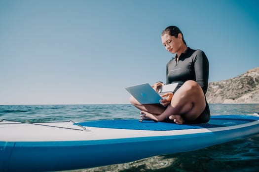 Woman sea laptop. Digital nomad, freelancer with laptop working on sup board at calm sea beach. Happy smiling girl relieves stress from work. Freelance, digital nomad, travel and holidays concept