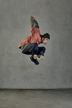 Stately man with tattooed body, beard. Dressed in hat, casual clothes and black sneakers. Jumping, dancing on gray background. Dancehall, hip-hop
