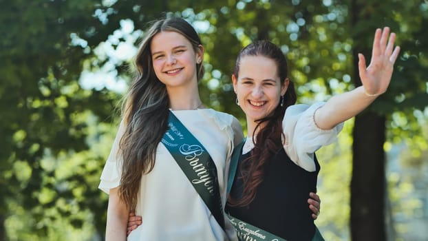 Two Russian schoolgirls graduate posing on a summer day with a ribbon and the name - Graduate 2023 on it.