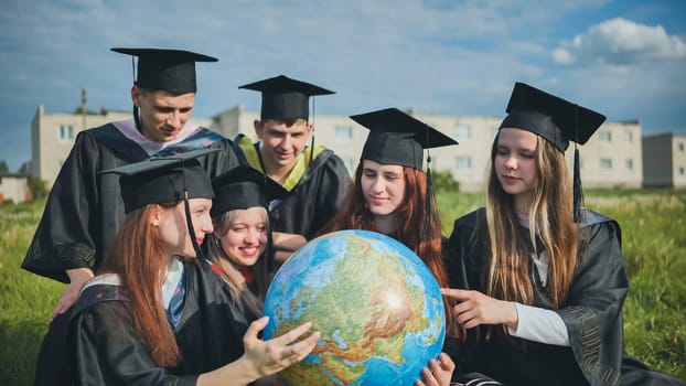 Graduates in black robes examine a geographical globe sitting on the grass.