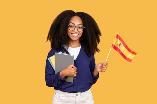 Happy black lady student with backpack and copybooks, holding Spanish flag, on yellow background