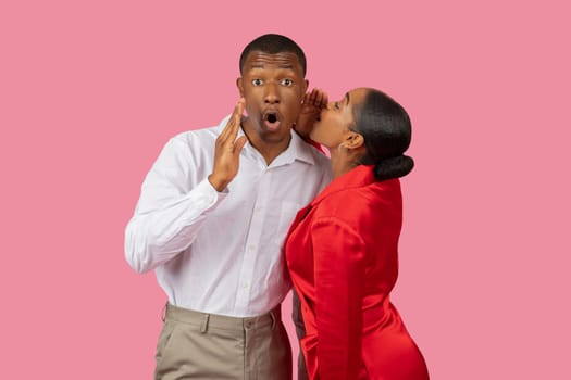 Black woman whispering to surprised man, both in elegant attire, pink background