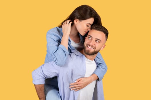 Woman whispers to man carrying her on yellow backdrop