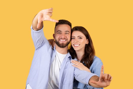 Happy couple making frame with hands, yellow backdrop