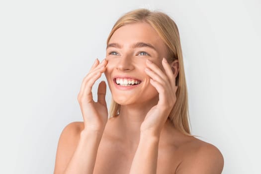 Cheerful blonde woman touching her soft moisturized face skin, studio