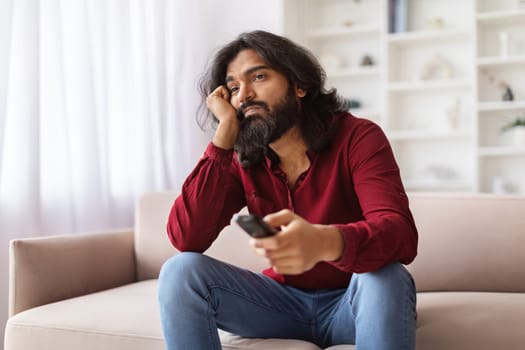 Unhappy eastern guy with remote control sitting on couch