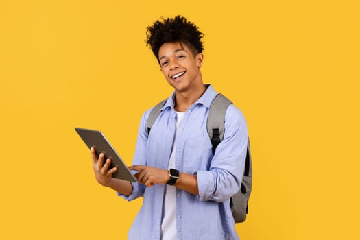 Black male student with tablet and backpack smiling on yellow
