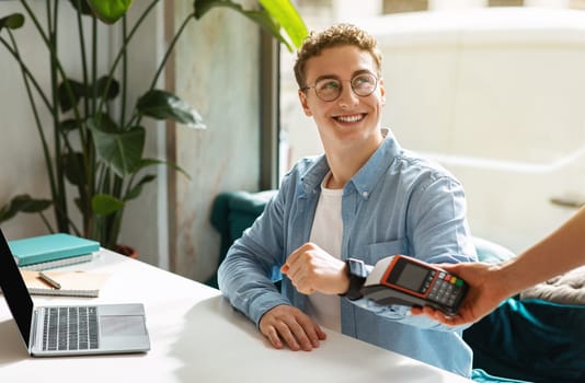 Happy young european guy using smartwatch for payment machine, sit on desk with laptop
