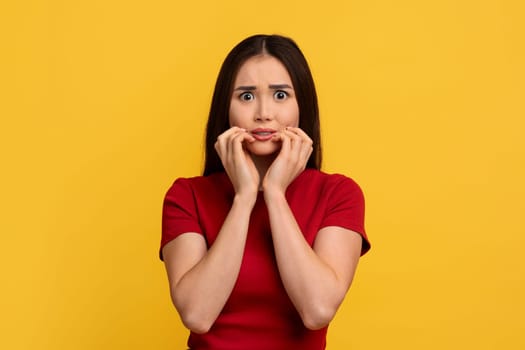Scared young asian woman touching her face, yellow wall background