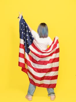 Victory Sign By Girl Covered With American Flag, Patriotic Pride, Cultural Diversity, And Freedom. Teenager Isolated On Yellow Backdrop With Copy Space For Your Text. Full-Height Backside View