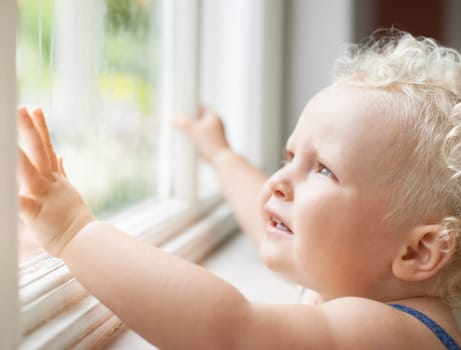 Baby, window and child thinking by glass at home, curious and sad alone. Young kid, blonde toddler and dream of cute girl at house, watching and childhood expression of learning with rain weather