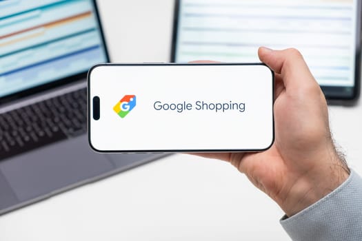 Google Shopping logo of app on the screen of mobile phone held by man in front of the laptop and tablet