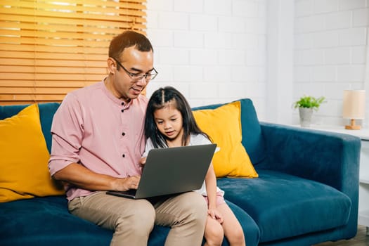 An Asian businessman finds happiness working from home on his laptop while his daughter uses a computer for e-learning. In their modern living room they share smiles laughter and quality family time.