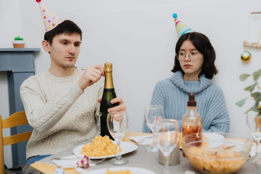 One young handsome Caucasian brunette with a cone hat on his head opens a bottle of champagne with a serious emotion, and a sad girl sits next to him at the table and celebrates his birthday, close-up side view.