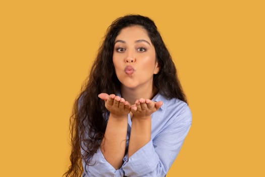Charming young woman with long curly hair blowing a kiss, hands cupped in front