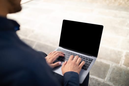 Cropped shot of unrecognizable young businessman typing on laptop with black empty screen, sitting outdoors in urban area, mockup for website advertisement and online business offers