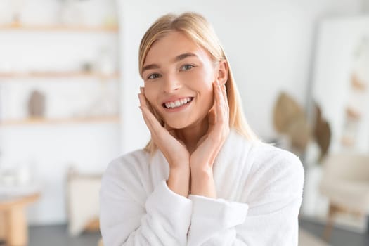 Blonde lady in bathrobe touches face showcasing glowing skin indoor