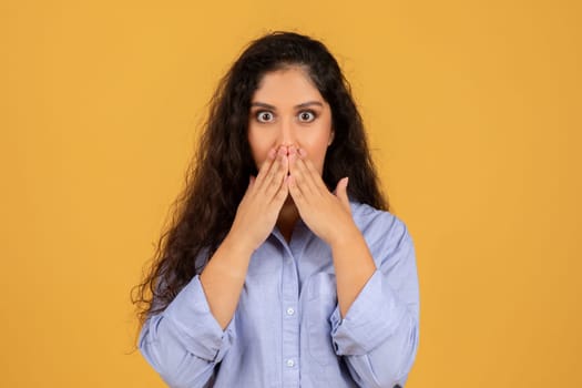 Startled young woman with voluminous curly hair covering her mouth with hands