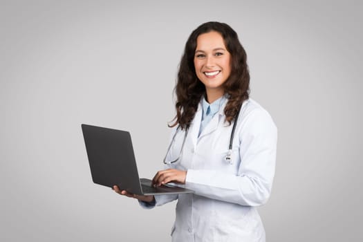 Online consultation service. Confident woman doctor using laptop, standing on grey studio background smiling at camera