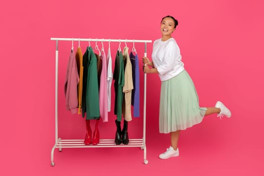 Smiling Asian lady standing beside garment rack filled with trendy clothes