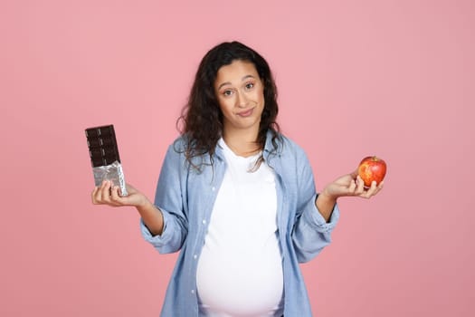 Confused young pregnant woman holding apple and chocolate bar