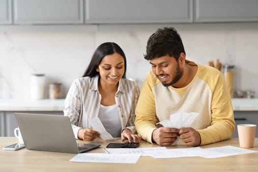 Smiling indian couple holding papers, calculating domestic bills, kitchen interior