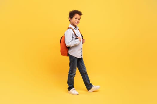 Black schoolboy walking with backpack on yellow background