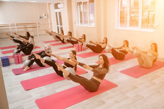 A group of six athletic women doing pilates or yoga on pink mats in front of a window in a beige loft studio interior. Teamwork, good mood and healthy lifestyle concept.