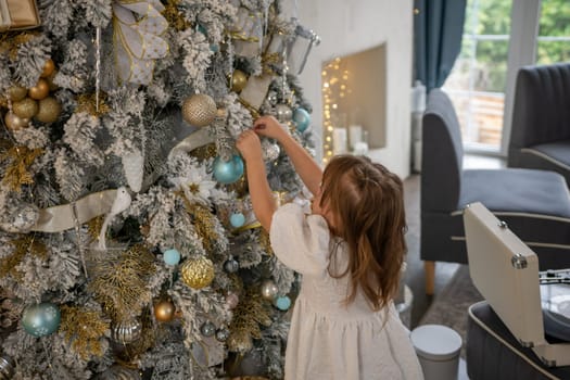A girl in a white dress decorates a Christmas tree with her own hands, Christmas holidays at home.