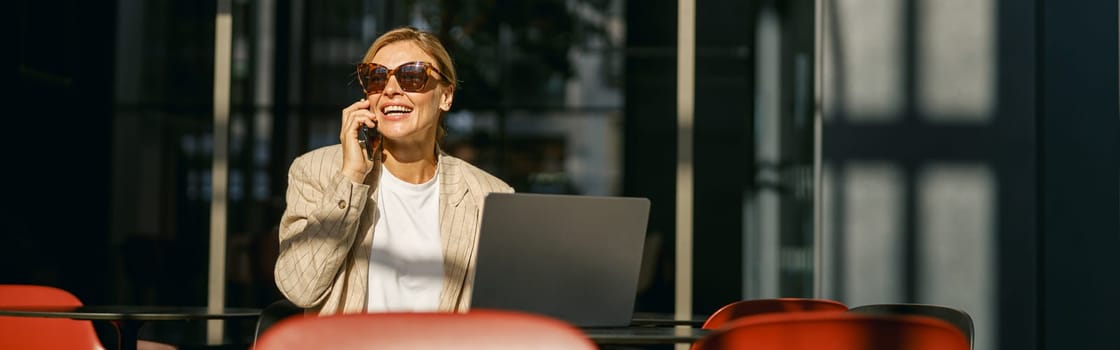Woman manager in sunglasses talking on phone with client while working on laptop in modern coworking