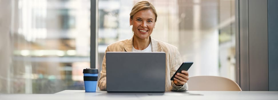 Cheerful female manager use phone while sitting in modern coworking and working on laptop