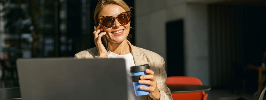 Businesswoman in sunglasses talking on phone with client while working on laptop in office