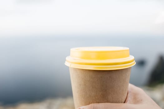 Yellow cup with lid, coffee against a backdrop of a blue sky and sea. Illustrating cup and beverage
