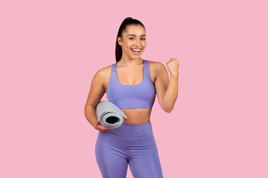 Happy caucasian woman in sportswear holding fitness mat and raising clenched fist up, celebrating win and triumph