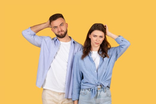 Confused couple with hands on head, puzzled expressions on yellow backdrop