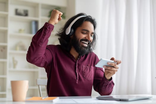 Overjoyed indian man wearing headphones celebrating win on his smartphone